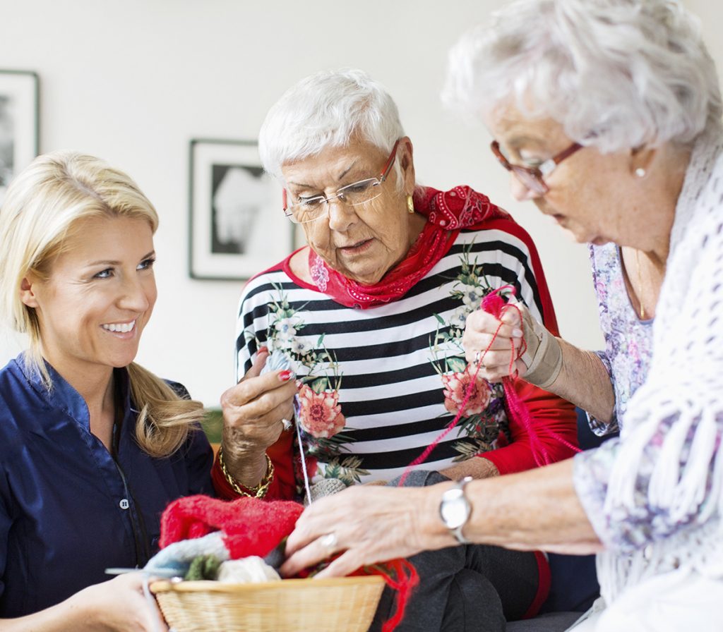 Happy female caretaker looking at senior women choosing wool for knitting at nursing home