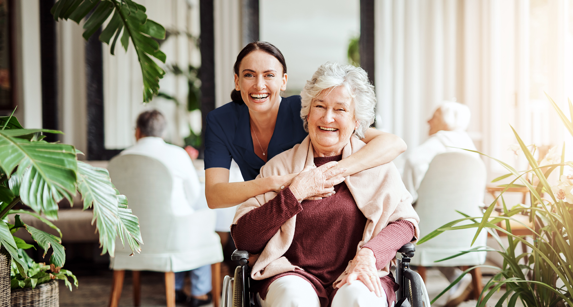 Plena employee putting her arms around an older woman in a wheelchair, both are smiling to the camera