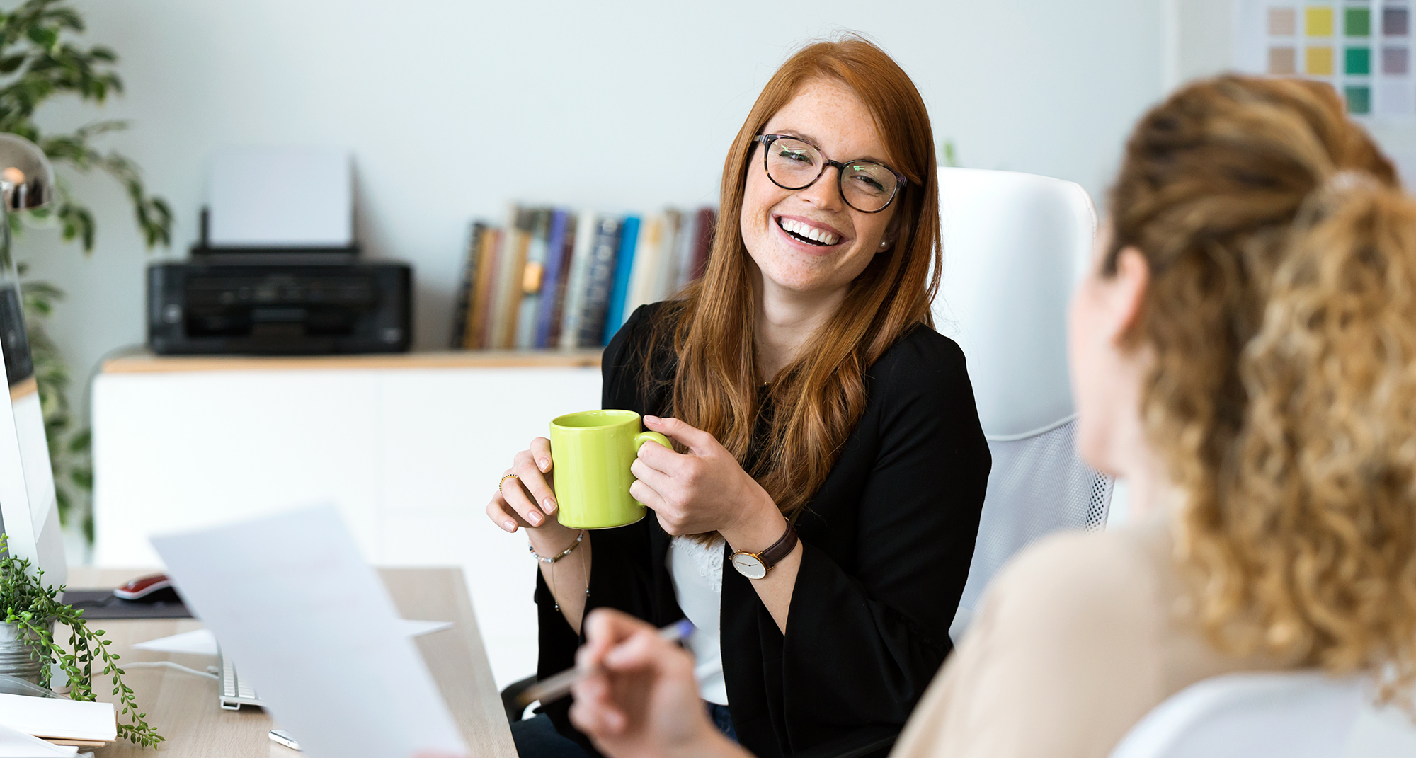 A woman with glasses holding a mug, laughing with her co-worker