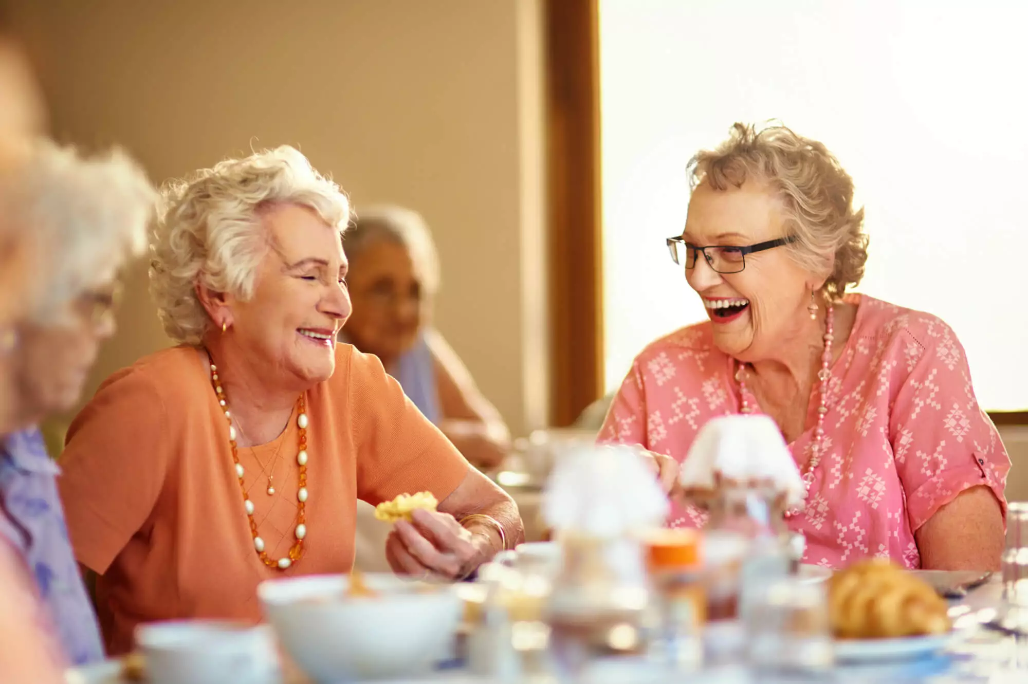 Two older women, in orange and pink, eating and laughing together