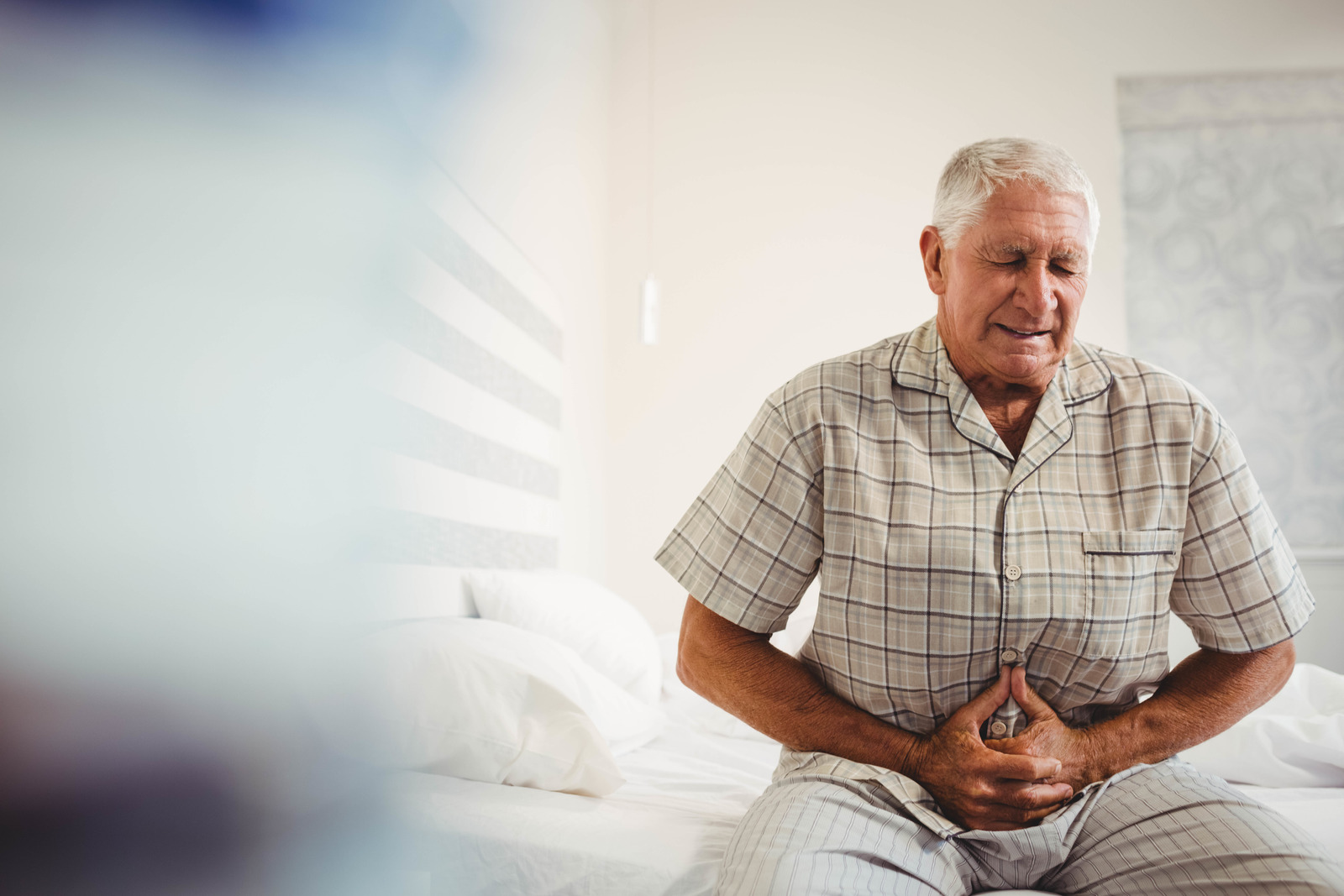 An older man in pain sitting on his bed while holding his stomach