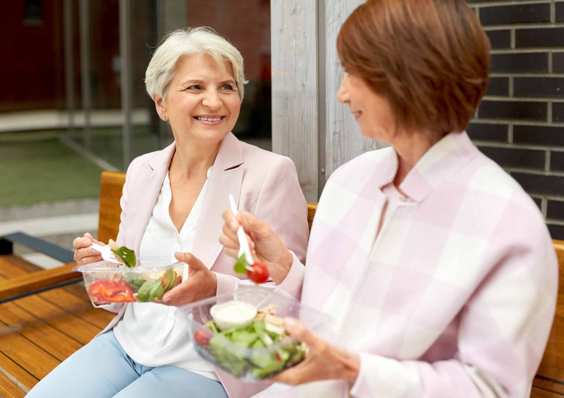 Two older white women sit on a bench outside eating salads from clear takeaway containers.