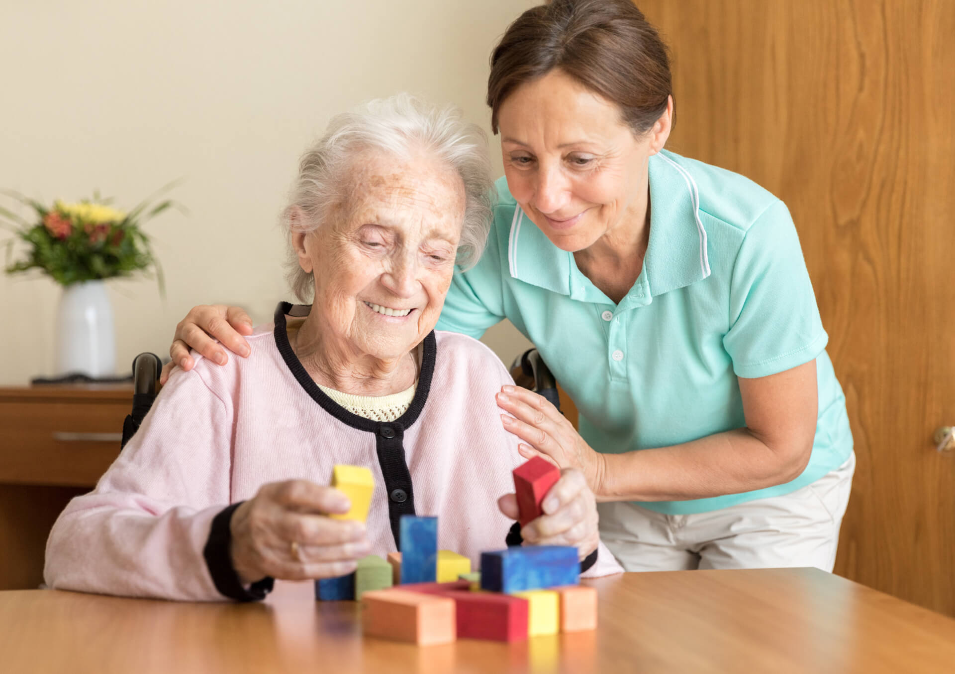 A woman in a polo top has her hand on the shoulders of an elderly woman who is holding coloured blocks. Both are smiling.