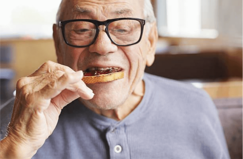 An elderly white man wearing glasses takes a bite of toast with jam on it.