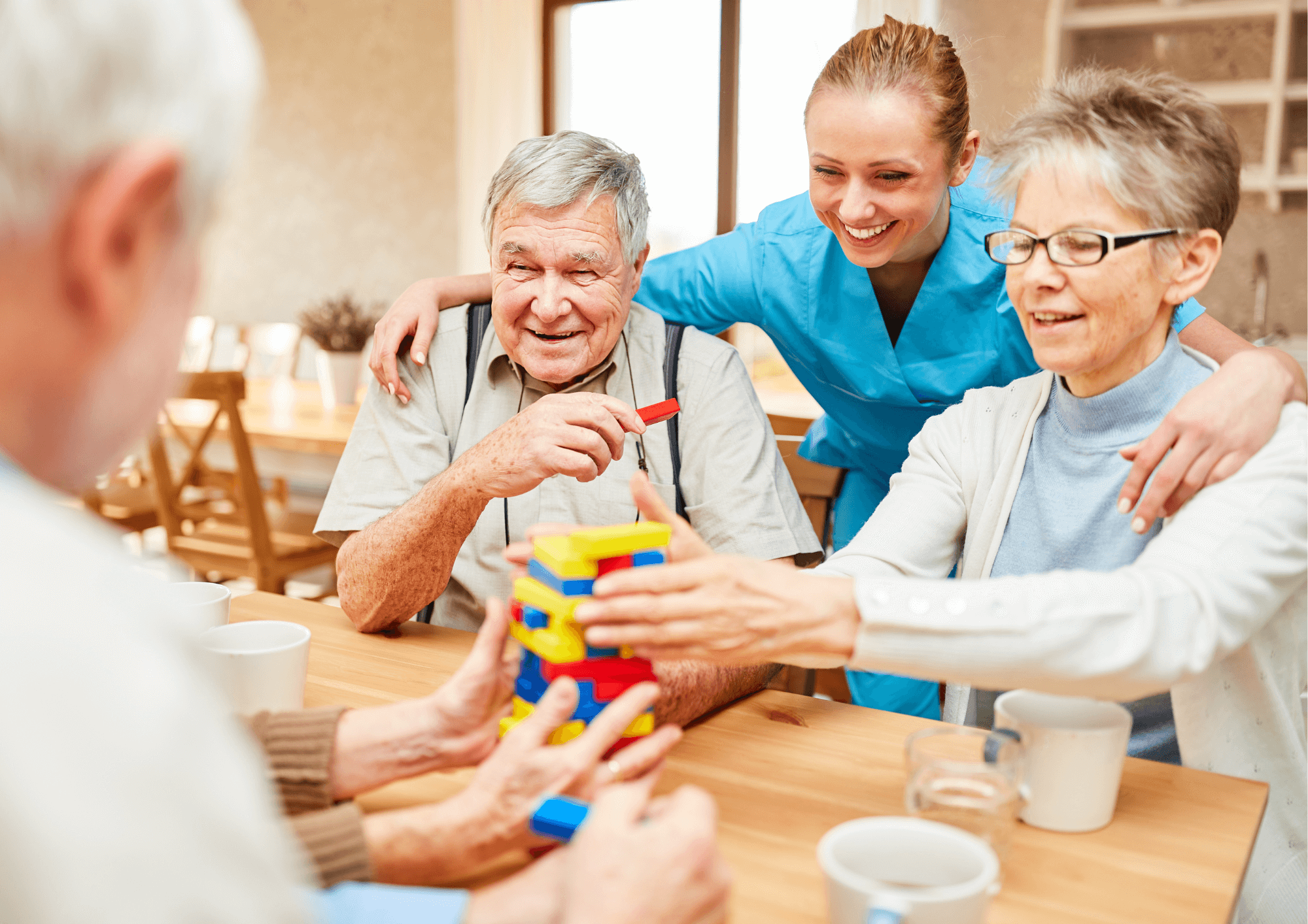 A group of elderly people sit at a table playing jenga. A younger female worker is standing with her arms around two of them.