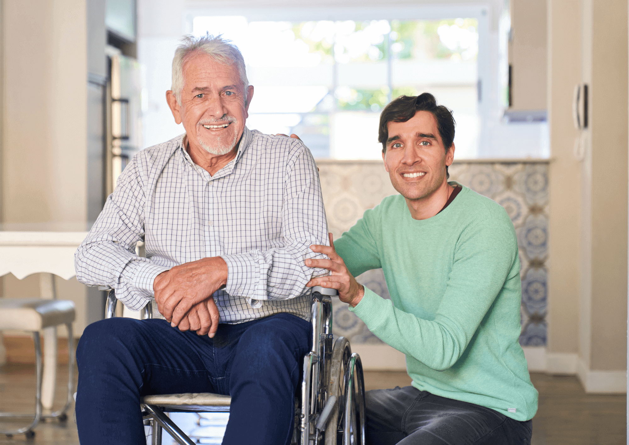 A younger man kneels by an older man in a wheelchair. They both smile at the camera.