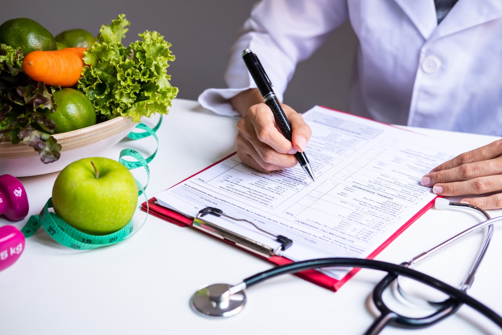 A Dietitian writing on a paper clipped to a board that’s placed on a table, the table has fruits, vegetables measuring tape on it.