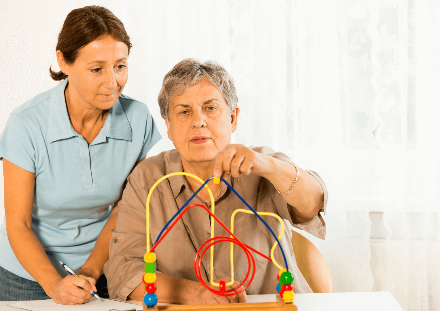 A woman stands behind an elderly woman who is moving blocks along a toy.